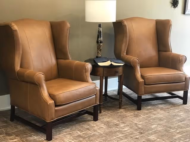 Two brown leather wingback chairs positioned on a carpeted floor with a small round wooden side table between them. The table holds a lamp and two open books. The background features a beige wall with a small decorative item partially visible.