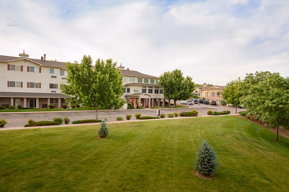 Front exterior of a senior living building with a manicured lawn, trees, driveway, and main entrance.