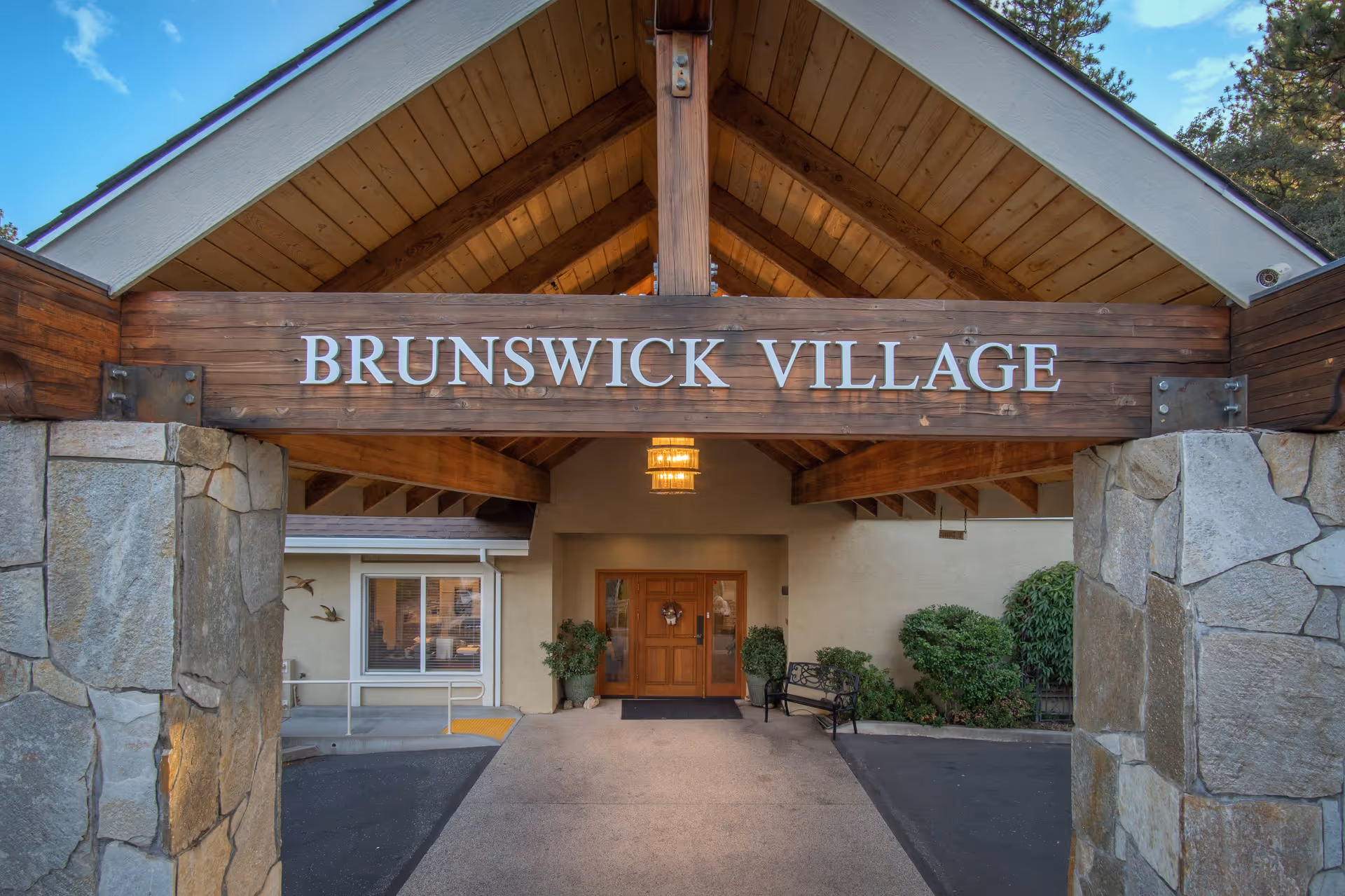 Entrance to Brunswick Village Assisted Living facility featuring a wooden sign with the facility name, stone pillars, a covered walkway with wooden beams, a bench, potted plants, and a double wooden door under a hanging light fixture.