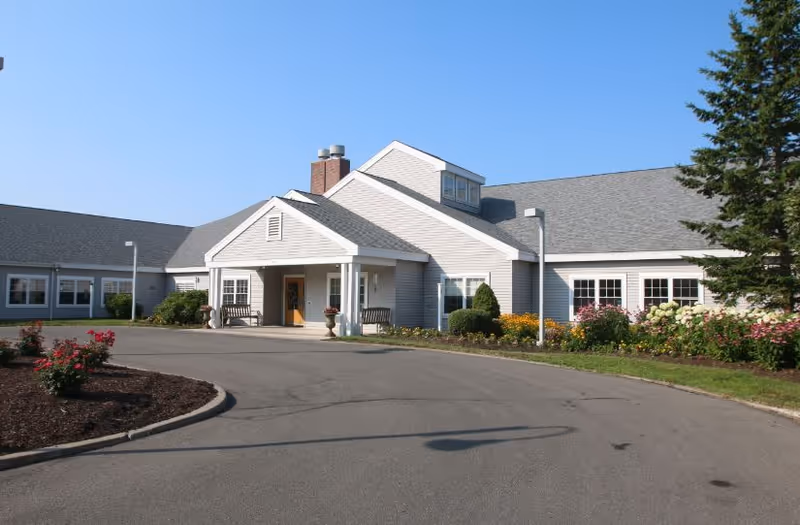 Exterior view of a single-story senior living facility building with a covered entrance, benches, and landscaped flower beds under a clear blue sky.