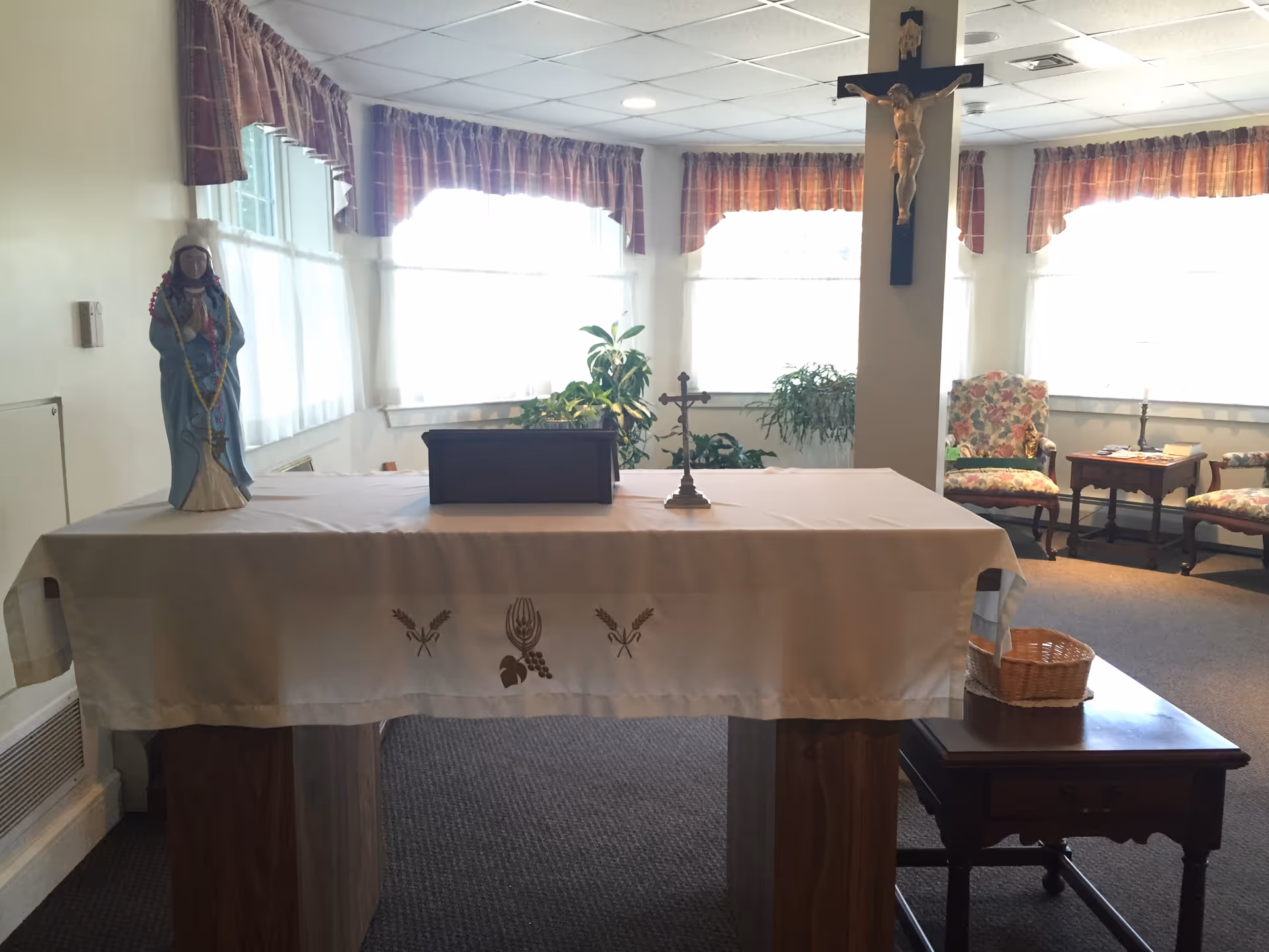 Small chapel-like interior with an altar draped in a cloth, a crucifix on a pillar, a statue, and chairs by bright windows.