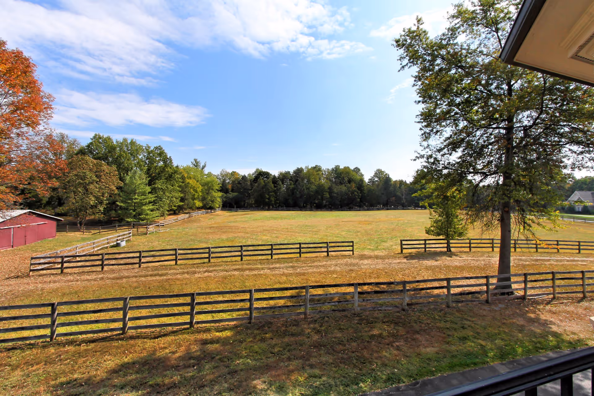 View of a large open grassy field surrounded by wooden fences with trees in the background under a partly cloudy blue sky, seen from a balcony or porch.