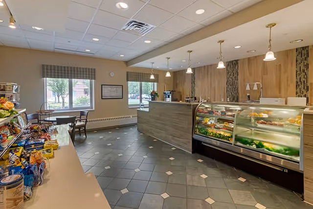 Interior view of a small café or snack bar area with a display case containing various food items, a counter, shelves stocked with snacks, and tables with chairs near windows with plaid valances.