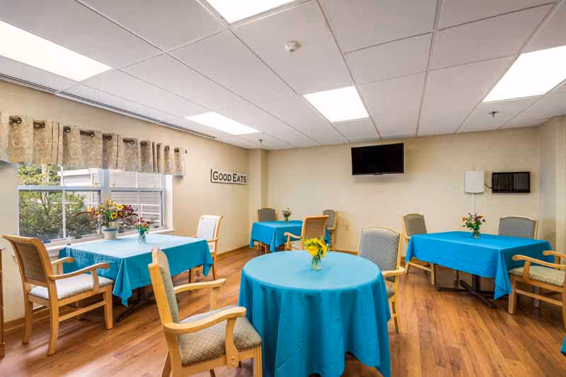 A bright dining room with several tables covered in blue tablecloths, each adorned with small flower arrangements. Wooden chairs with cushions surround the tables. A window with patterned curtains lets in natural light, and a wall-mounted TV is visible. A sign on the wall reads 'GOOD EATS'.