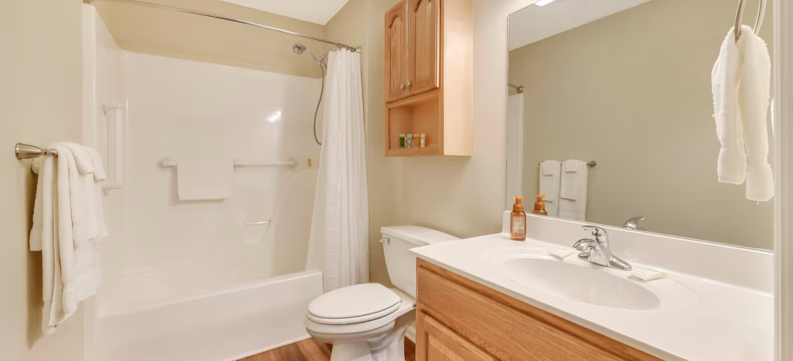 A clean and well-lit bathroom featuring a white bathtub with a shower curtain, a toilet, a wooden vanity with a sink, and a large mirror above the sink. White towels hang on towel racks near the bathtub and beside the sink. A wooden cabinet is mounted on the wall above the toilet.