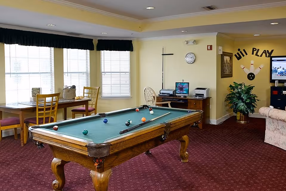 Bright common room with a pool table in the center, a table and chairs by windows, a desk with computers, and a TV on yellow walls.