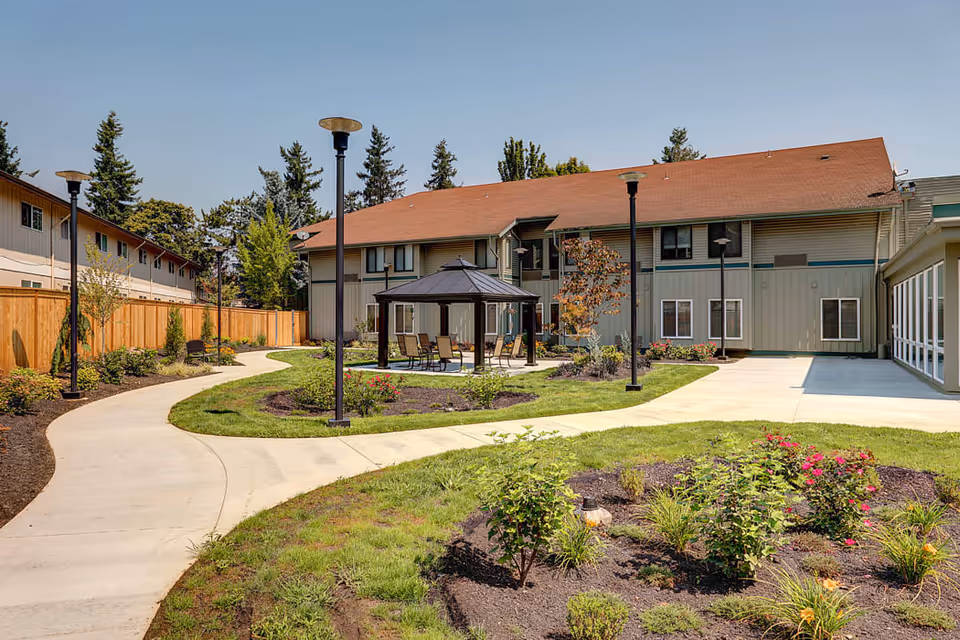 Outdoor garden area at Firwood Gardens featuring a paved walkway, landscaped flower beds, a gazebo with chairs underneath, surrounded by a two-story building and tall trees in the background under a clear sky.