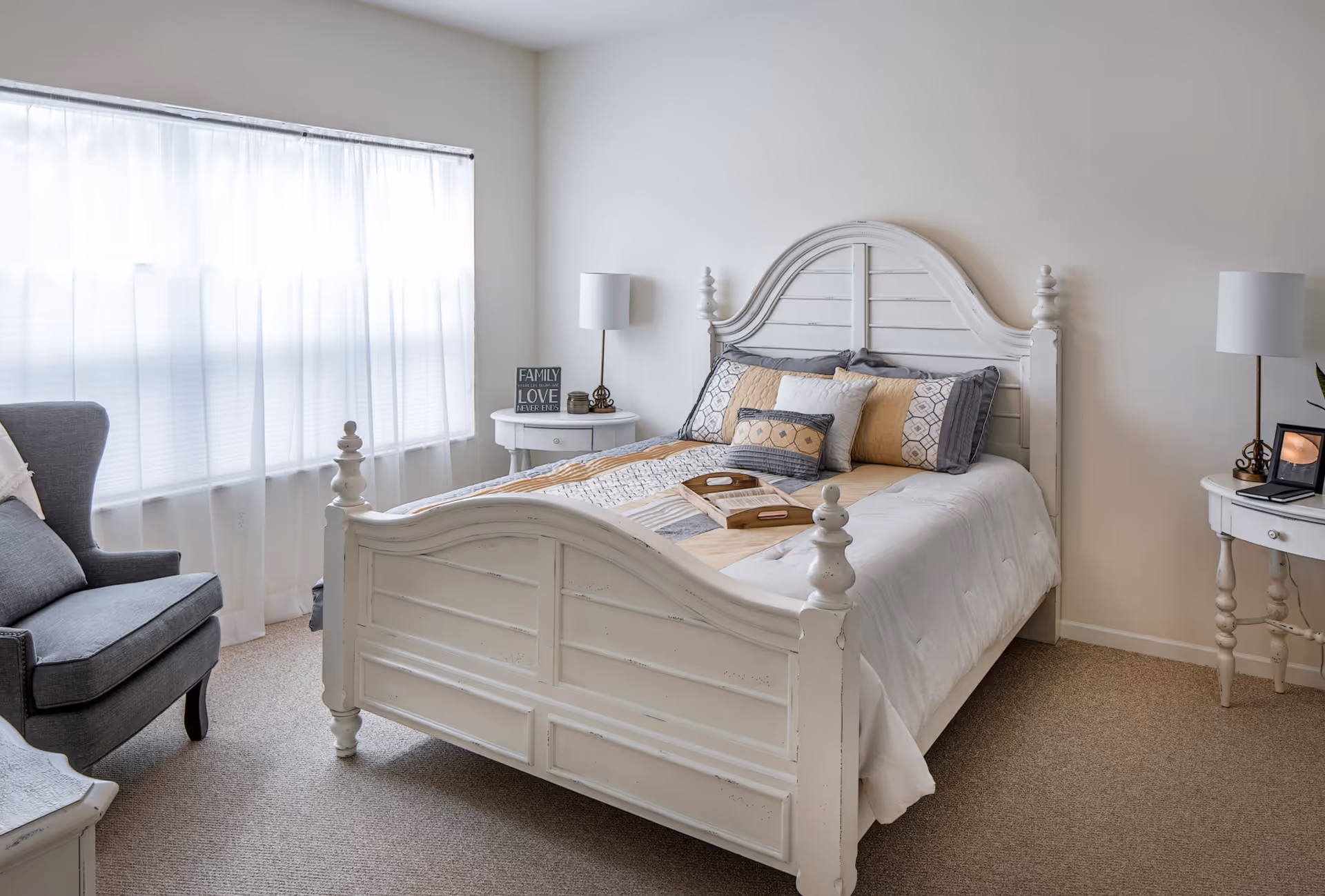 A bright bedroom with a white wooden bed frame and a neatly made bed with patterned pillows and a quilt. There are two white nightstands on either side of the bed, each with a lamp. A gray armchair is positioned near a large window with sheer white curtains, allowing natural light to fill the room.