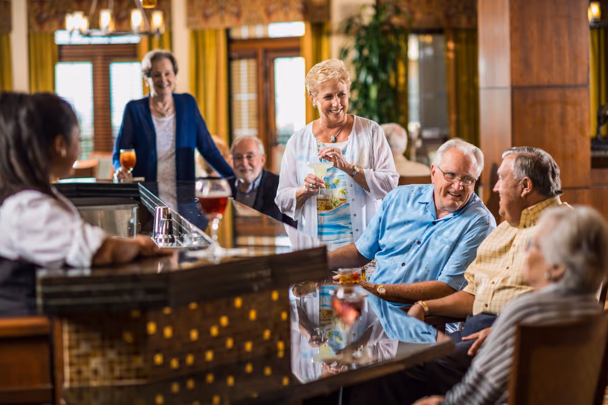 A group of elderly people socializing at a bar inside a senior living facility. One woman stands holding a cocktail, while others sit around the bar counter engaged in conversation. A bartender is seen on the left side of the image. The setting is warm and inviting with wooden decor and large windows with yellow curtains in the background.