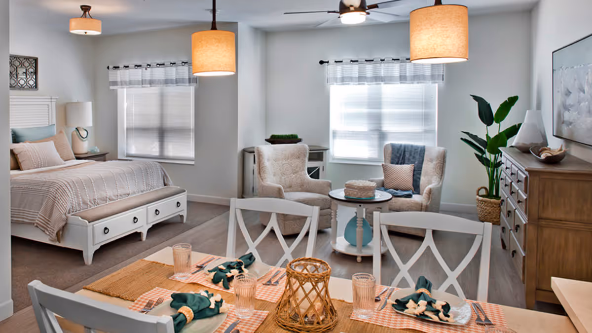 A bright and cozy senior living suite at Trinity Springs featuring a bedroom area with a white bed, beige bedding, and a bench at the foot of the bed. Adjacent to the bedroom is a sitting area with two upholstered armchairs and a small round table. In the foreground, there is a dining table set with placemats, napkins, glasses, and cutlery. The room has large windows with blinds and curtains, ceiling lights, a ceiling fan, a wooden dresser, and a potted plant.