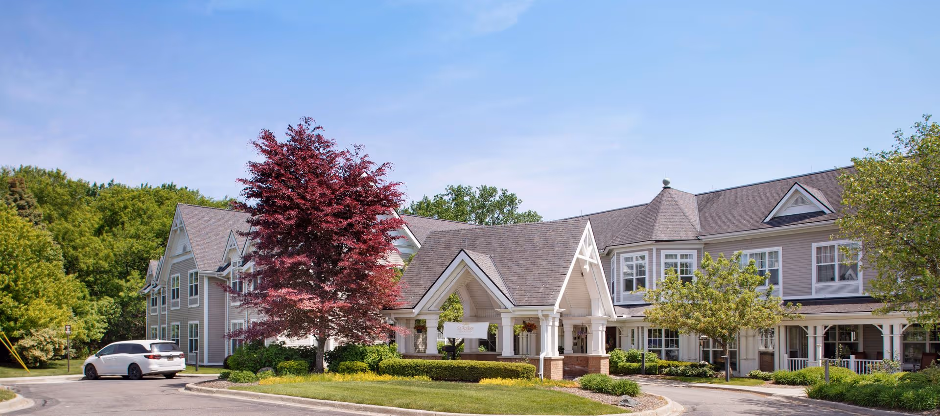 Exterior view of a senior living facility named Sunrise of West Bloomfield, featuring a large building with multiple windows, a covered entrance, well-maintained landscaping, green trees, and a clear blue sky.
