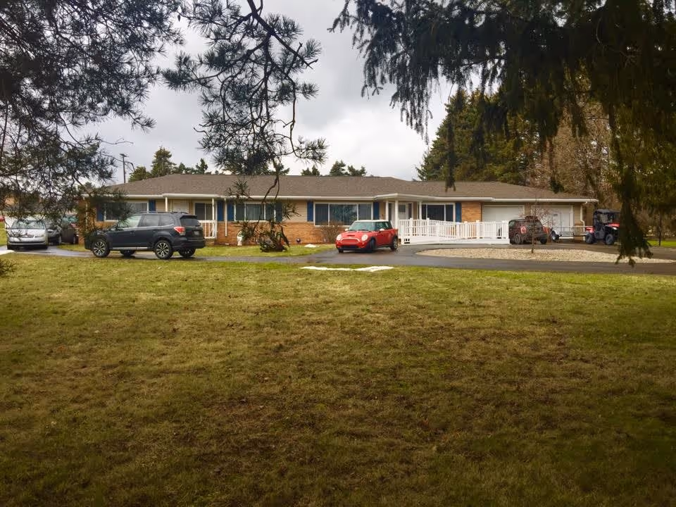 Single-story brick building with several windows and a white ramp entrance, surrounded by a grassy lawn and trees. Multiple cars are parked in front of the building under a cloudy sky.