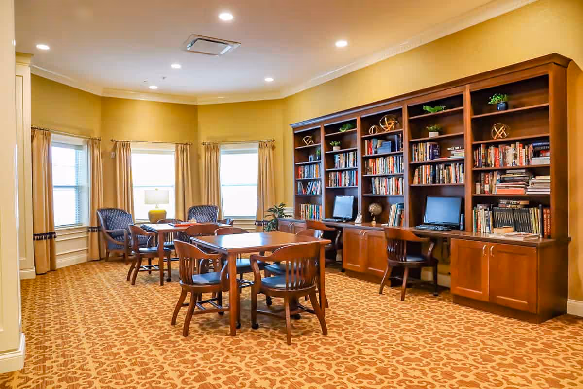 A cozy library or common area with wooden bookshelves filled with books and decorative items, two computer workstations, several wooden tables with chairs, and two armchairs near windows with beige curtains. The room has warm yellow walls and patterned carpet flooring.