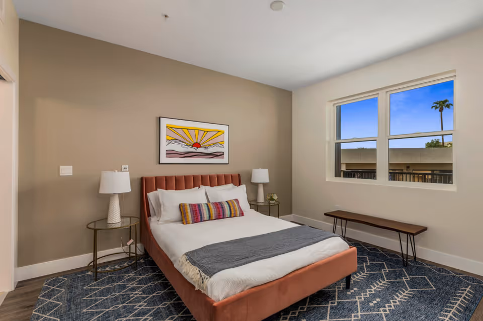 A furnished bedroom with a bed, bedside tables and lamps, wall art, a window, and a bench.