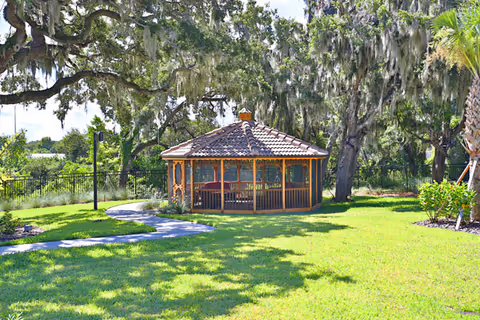 A wooden gazebo with a tiled roof situated in a lush green garden area with a winding concrete pathway leading to it. Large trees with hanging moss surround the gazebo, and there are landscaped plants and a palm tree nearby under a bright sky.