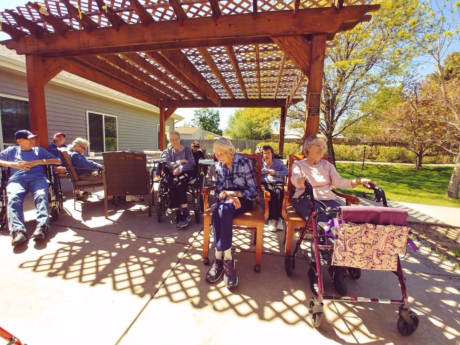Elderly residents seated under a wooden pergola on a sunny patio, some using walkers and wheelchairs.