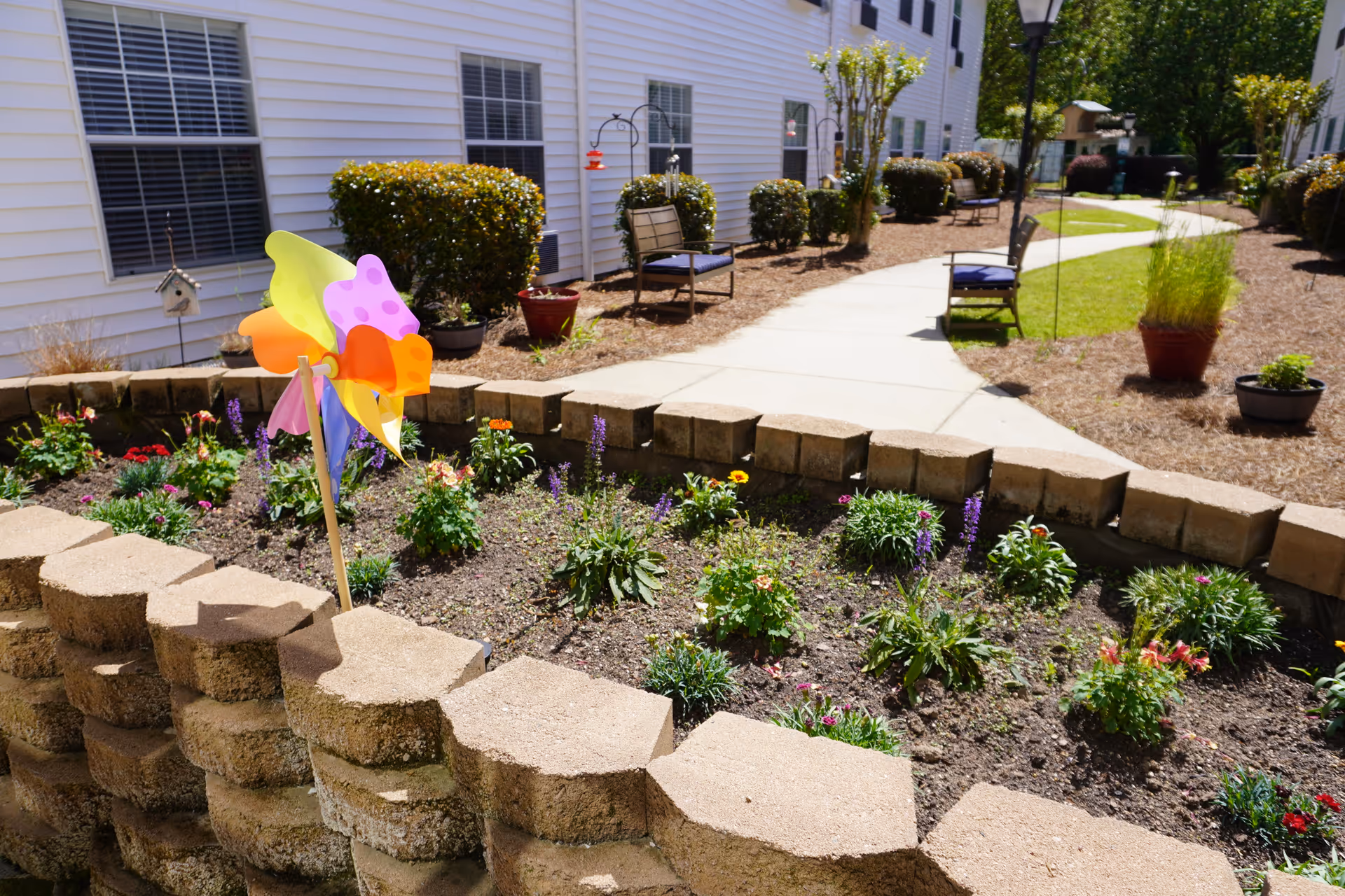Outdoor garden area at Sodalis Martinez with a raised flower bed containing various blooming plants and a colorful pinwheel. A paved walkway curves through the landscaped area with benches and shrubs alongside a white building.