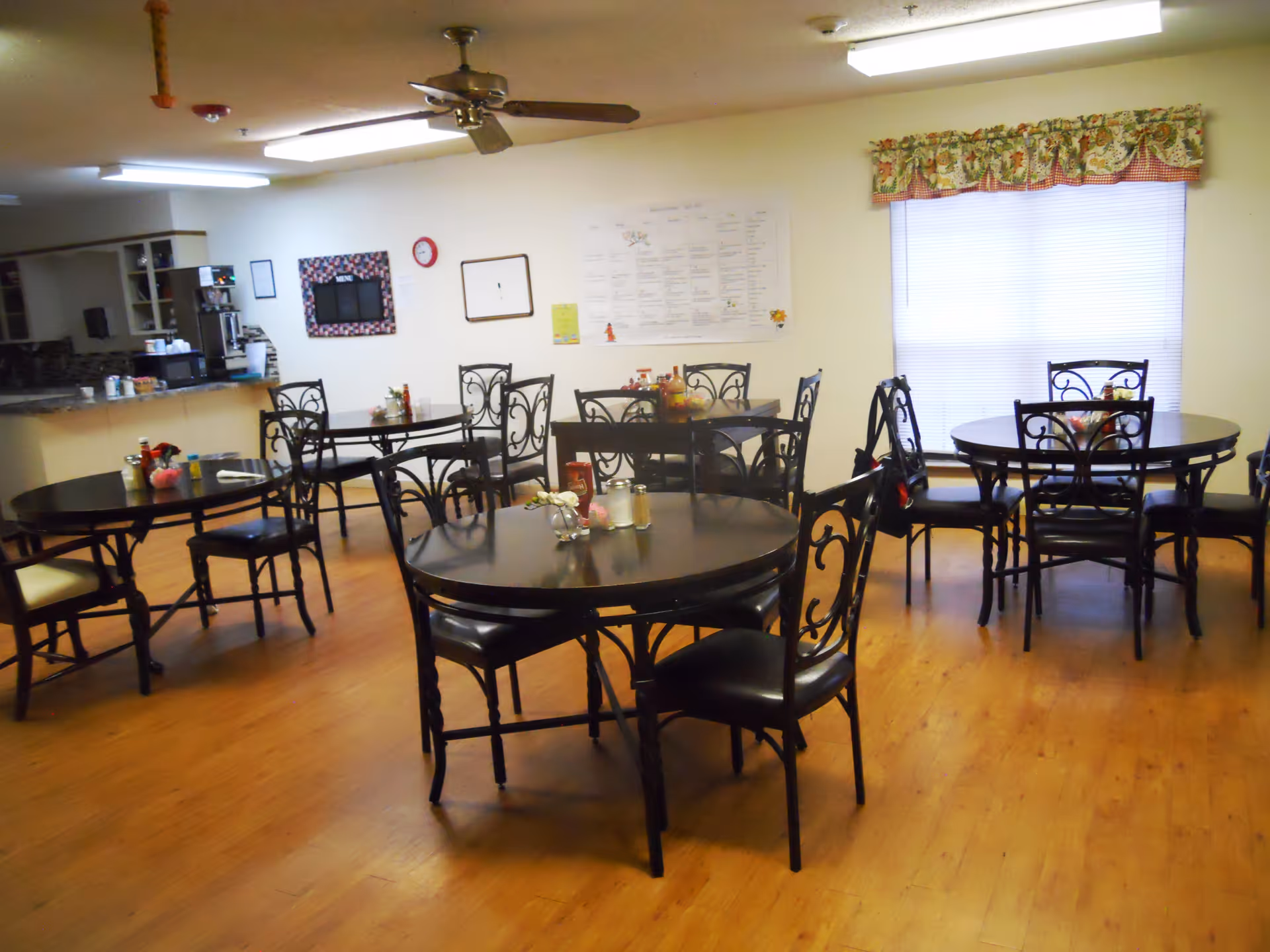 Dining area with several round tables and black metal chairs with cushions. The room has wooden flooring, a ceiling fan, and a window with floral valance curtains. A kitchen counter is visible in the background with appliances and a bulletin board on the wall.