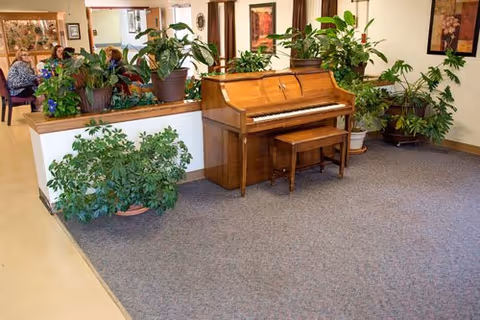 Interior view of a senior living facility common area featuring a wooden upright piano with a matching bench surrounded by various green potted plants. In the background, there are people seated at tables, and the walls are decorated with framed artwork.