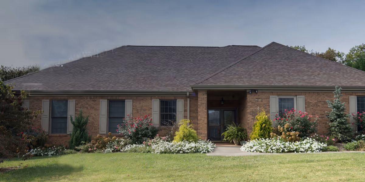 Front exterior view of a single-story brick building with a dark shingled roof, several windows with shutters, a covered entrance with double doors, and well-maintained landscaping including green grass, bushes, and flowering plants.