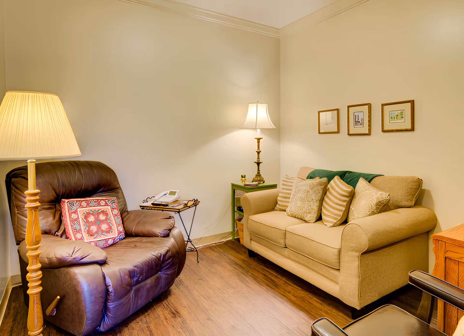 A cozy living room with a brown leather recliner chair featuring a decorative pillow, a beige sofa with multiple patterned cushions, two table lamps providing warm lighting, a small green side table with items on it, and three framed pictures hanging on the wall above the sofa. The room has wooden flooring and light-colored walls.