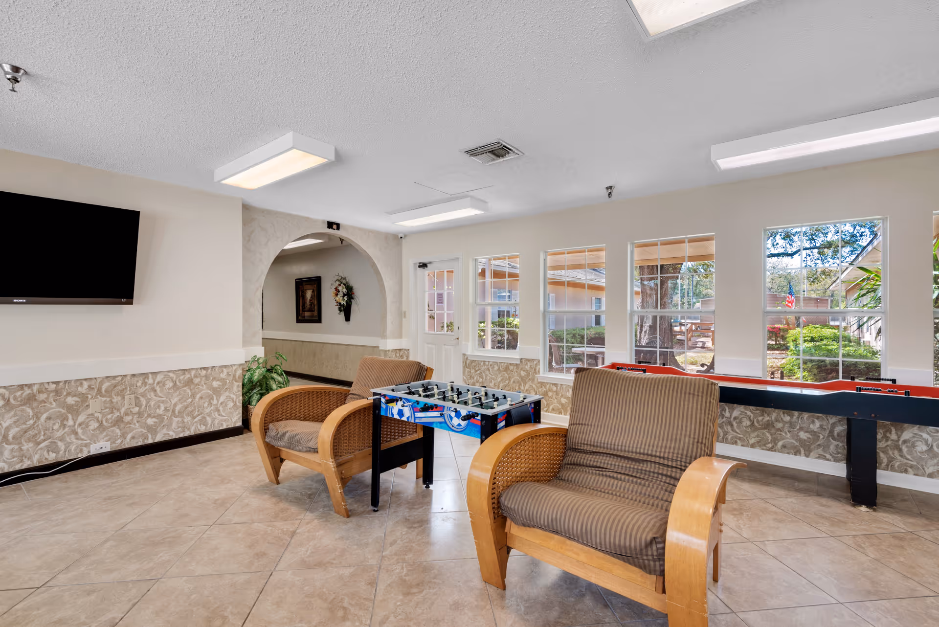 A bright common room in a senior living facility with two cushioned armchairs, a foosball table, and a shuffleboard table near windows showing an outdoor garden area. A flat-screen TV is mounted on the wall, and there is an arched doorway leading to another room.