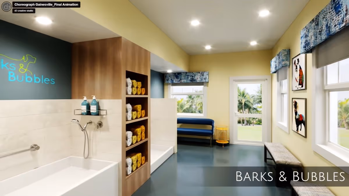 Interior view of a pet grooming area named Barks & Bubbles featuring two white pet bathing tubs with mounted shower heads and soap dispensers. Between the tubs is a wooden shelving unit stocked with rolled yellow and white towels. The room has light yellow walls, blue flooring, and windows with blue patterned valances. There is a blue bench and a small yellow side table near a glass door leading outside. Two colorful dog paintings hang on the wall above a cushioned bench.