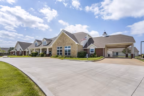 Exterior view of a single-story senior living facility building with a stone and brick facade, multiple windows, a covered entrance, an American flag on a flagpole, and a wide concrete driveway under a partly cloudy sky.