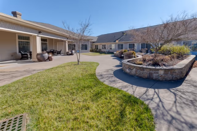 Outdoor courtyard area at Cedar Creek of Bloomington Memory Care featuring a curved concrete walkway, green grass, leafless trees, large stone planter beds, and a covered patio with seating and large decorative pots.