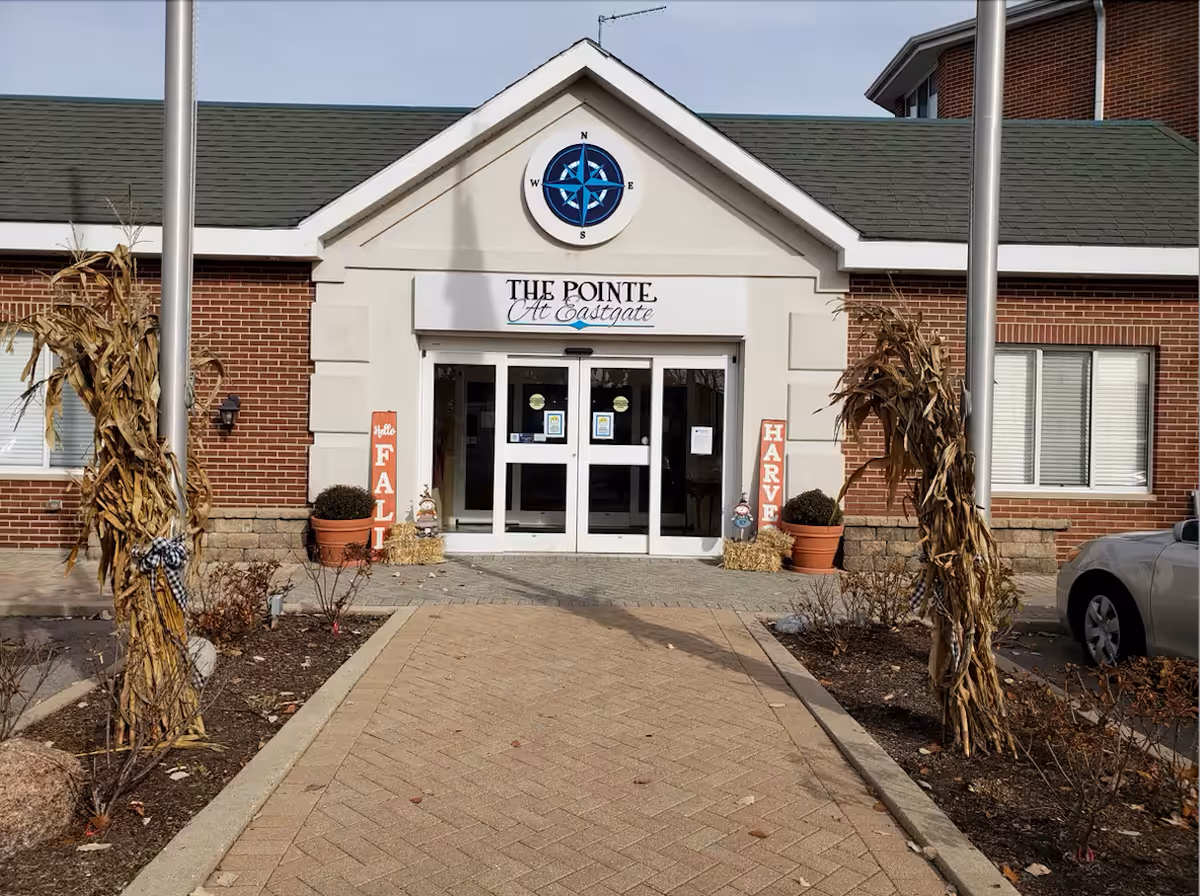 Entrance to The Pointe at Eastgate facility with automatic sliding glass doors, a compass emblem above the entrance, fall decorations including cornstalks tied to flagpoles, potted plants, hay bales, and signs that say 'Hello Fall' and 'Harvest'.