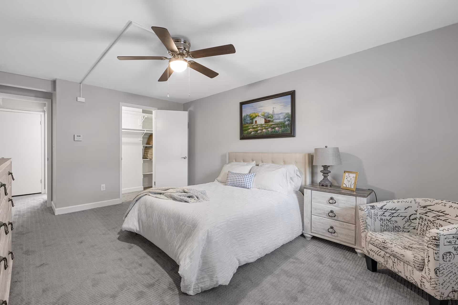 A neatly arranged bedroom with a bed covered in white bedding, a nightstand with a lamp and framed picture, a patterned armchair, a dresser, and an open closet door. The walls are painted light gray and there is a ceiling fan with a light fixture. A framed painting hangs above the bed.