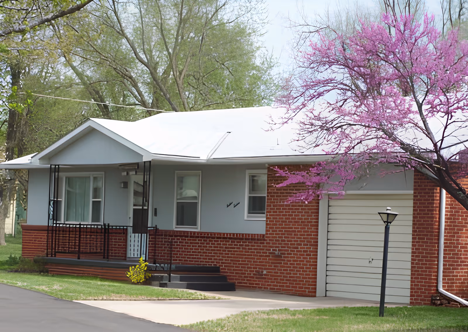 Exterior view of a single-story brick and light gray house with a white roof, a small front porch with black railings, and a garage door. There is a blooming tree with pink flowers on the right side and green trees in the background.