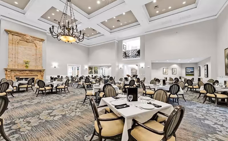 A spacious dining room with multiple tables covered in white tablecloths, each set with plates, glasses, and silverware. The room features high ceilings with recessed lighting and a large decorative chandelier. There is a large ornate fireplace on one wall and several framed artworks on the walls. The carpet has a floral pattern, and the chairs have striped upholstery.