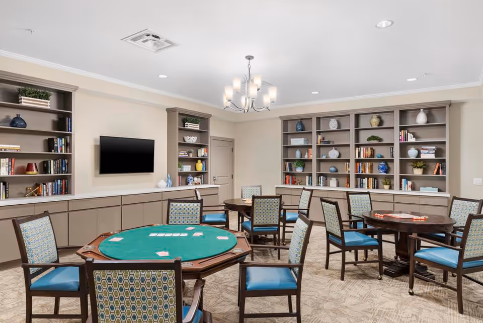 A well-lit game room with multiple round tables and chairs arranged around them. One table has a green felt surface with playing cards on it, and another table has a board game set up. The room features built-in bookshelves filled with books, decorative vases, and plants. A flat-screen TV is mounted on the wall above cabinets, and a modern chandelier hangs from the ceiling.