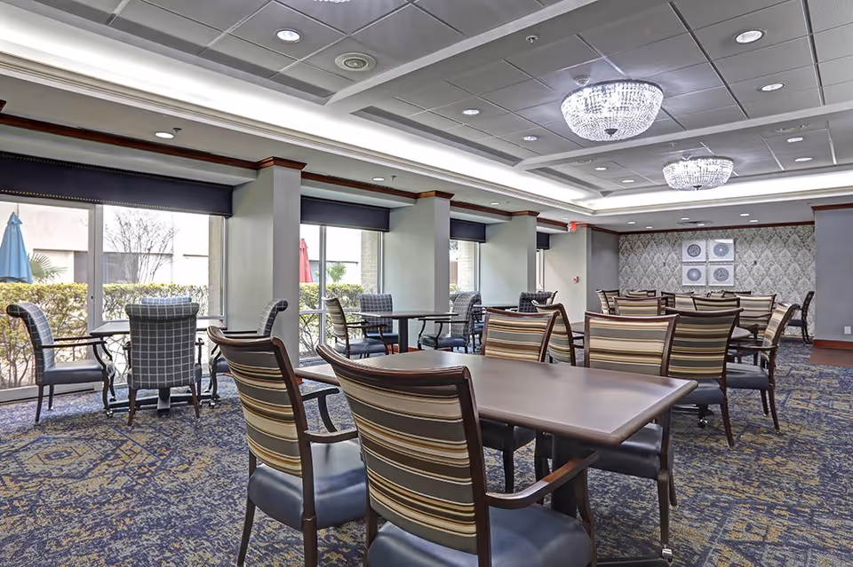 A spacious dining room with multiple tables and chairs arranged neatly. The room features large windows with black valances, allowing natural light to enter. The ceiling has recessed lighting and two elegant chandeliers. The carpet has a patterned design, and the walls are decorated with framed artwork and wallpaper.