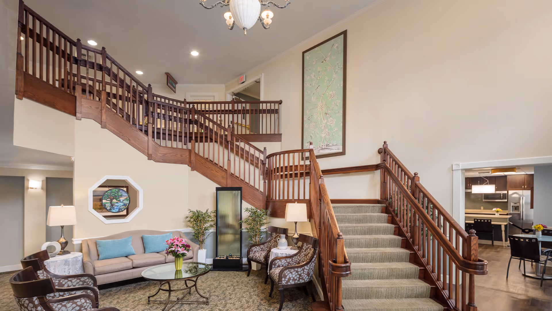 Interior view of a senior living facility lobby area featuring a wooden staircase with carpeted steps and wooden railings. The seating area includes a beige sofa with blue cushions, two patterned armchairs, a glass coffee table with a vase of flowers, and two side tables with lamps. There are decorative plants and artwork on the walls. In the background, a dining area and kitchen are partially visible.