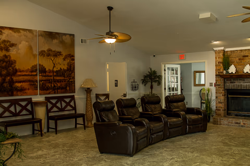 A cozy common area in a senior living facility featuring four dark brown leather recliners arranged in a row facing a brick fireplace. The room has a ceiling fan with a light, two wooden chairs with cross-back designs against the wall, a tall decorative lamp, and a large triptych painting of a nature scene with trees and a cloudy sky. There are plants near the fireplace and an exit sign above a doorway in the background.