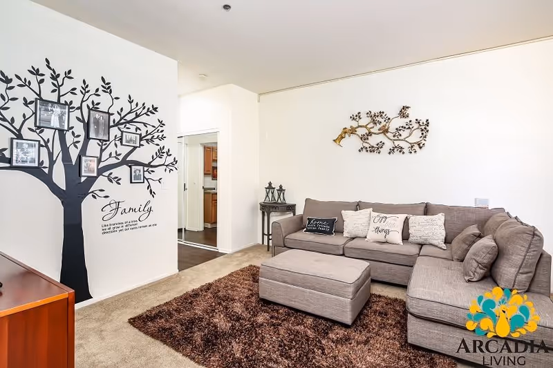 A cozy living room with a large gray sectional sofa adorned with decorative pillows, a matching ottoman, and a brown shaggy rug. On the wall above the sofa is a metal wall art piece shaped like a tree branch with birds. Another wall features a black tree decal with family photos hanging on its branches and the word 'Family' along with a quote beneath it. A small side table with lanterns is placed next to the sofa. The room has beige carpet and white walls, with a doorway leading to a kitchen area. The Arcadia Living logo is visible in the bottom right corner.