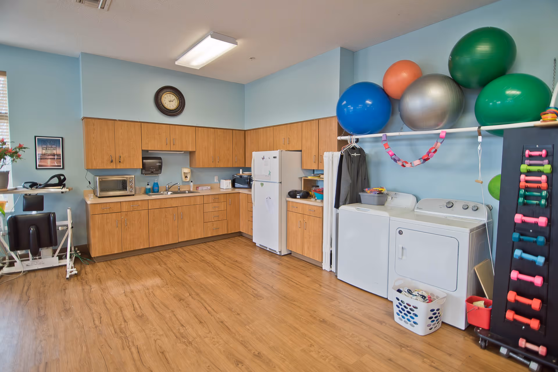 A multi-purpose room with a kitchenette featuring wooden cabinets, a microwave, a refrigerator, and a sink. On the right side, there is a washer and dryer with a laundry basket in front. Above the laundry machines, several large exercise balls are stored on a wall-mounted rack. To the right, a rack holds colorful dumbbells. The floor is wooden, and the walls are painted light blue. A clock is mounted above the cabinets, and some exercise equipment is visible on the left side near a window.