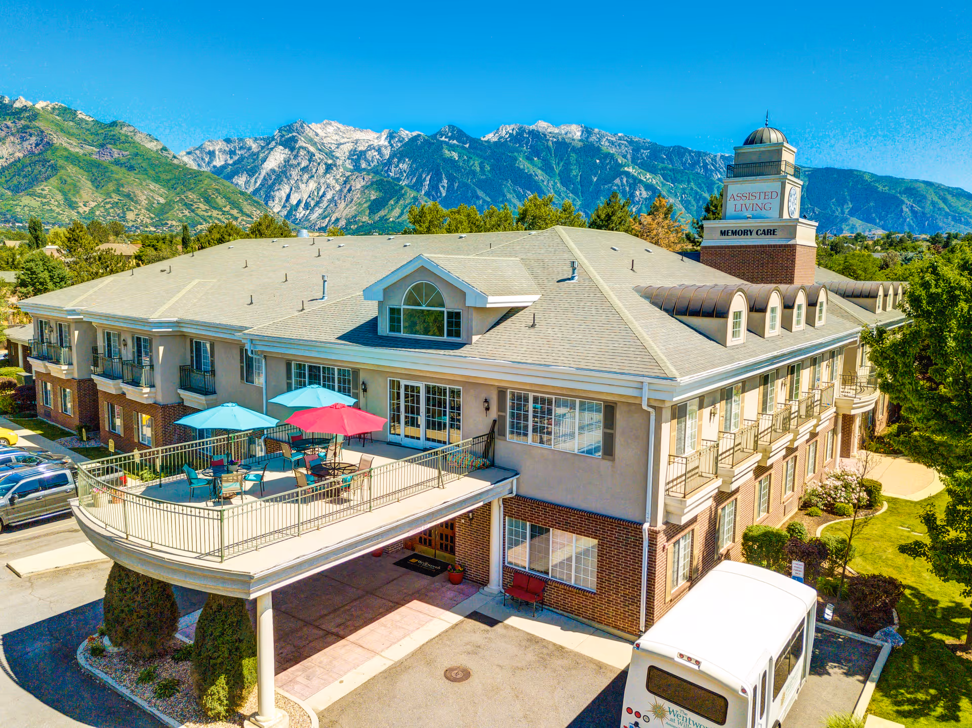 A large assisted living facility building with a covered entrance and an outdoor patio area featuring tables with colorful umbrellas. The building has multiple windows and balconies, and is set against a backdrop of tall, rugged mountains under a clear blue sky. A white shuttle bus is parked near the entrance.