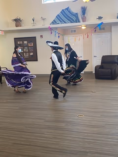 Three people wearing traditional Mexican dance costumes and face masks are performing a dance in a room with wooden flooring. The room has beige walls, a brown leather chair, a framed picture on the wall, and colorful decorations hanging from the ceiling.