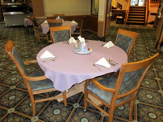 A round dining table covered with a lavender tablecloth set with four place settings, each with a folded white napkin, fork, knife, and spoon. The table has four wooden chairs with patterned upholstery. In the background, there are more tables and chairs, and a staircase leading to another level.