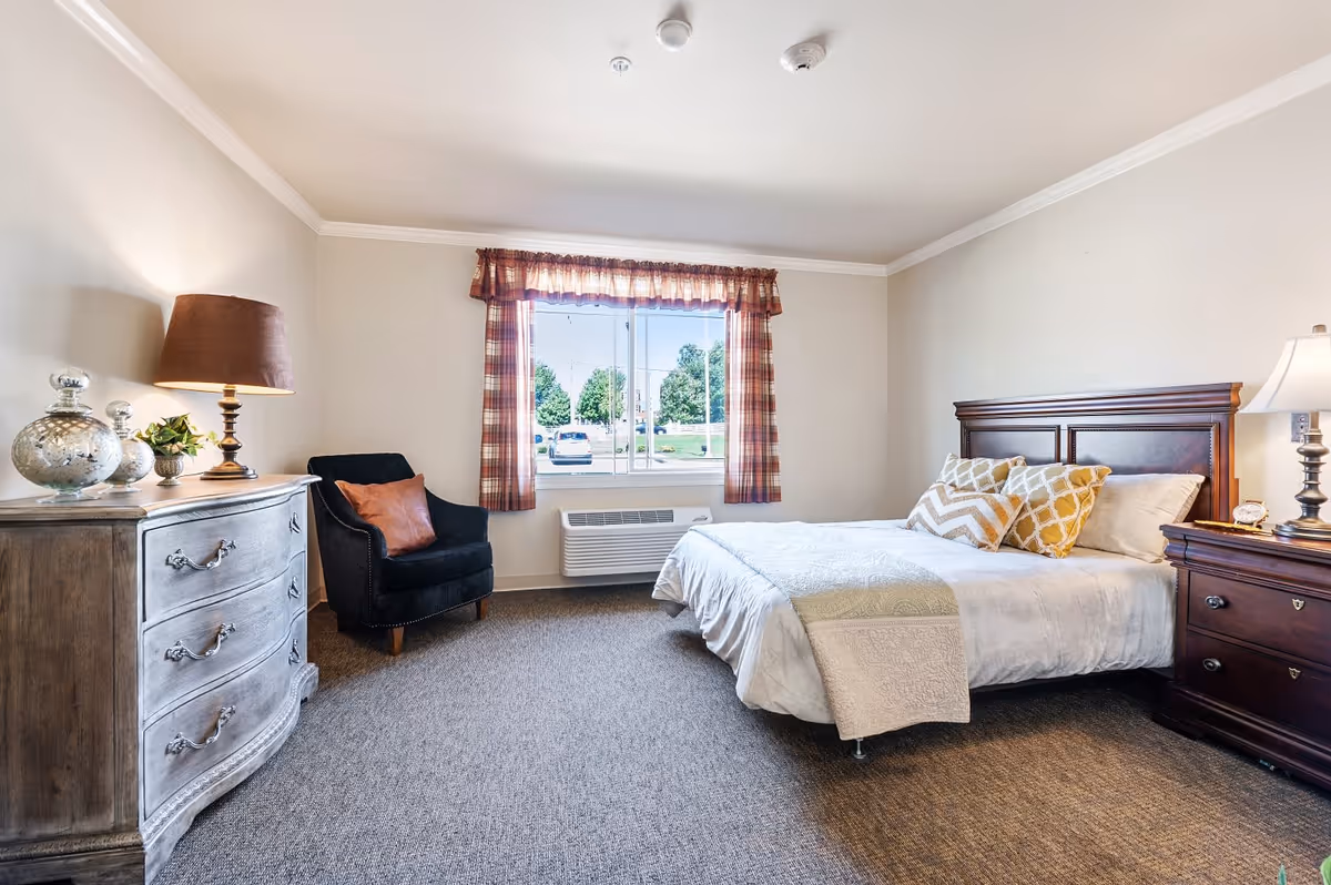 A bright and cozy bedroom with a large window covered by plaid curtains. The room features a comfortable bed with white bedding and decorative pillows, a dark wooden nightstand with a lamp, a vintage-style dresser with a lamp and decorative items, and a black armchair with a brown pillow. The carpeted floor and neutral wall colors create a warm and inviting atmosphere.