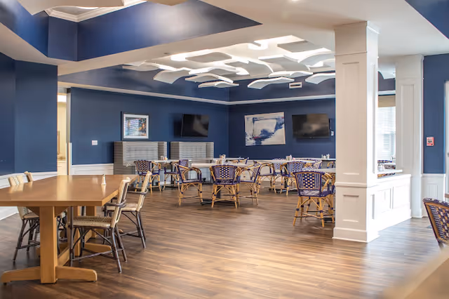 Spacious communal dining room with blue walls and wood flooring, arranged tables and wicker chairs, two wall-mounted TVs and sculptural white ceiling panels.