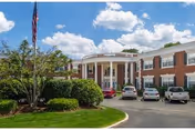 Front exterior of a two-story brick senior living facility with white columns, parked cars, landscaped shrubs, and an American flag under a blue sky.