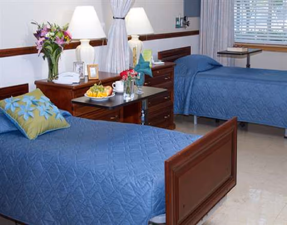 Two twin beds with blue quilts in a bedroom; lamps, flowers, and fruit on a surface.