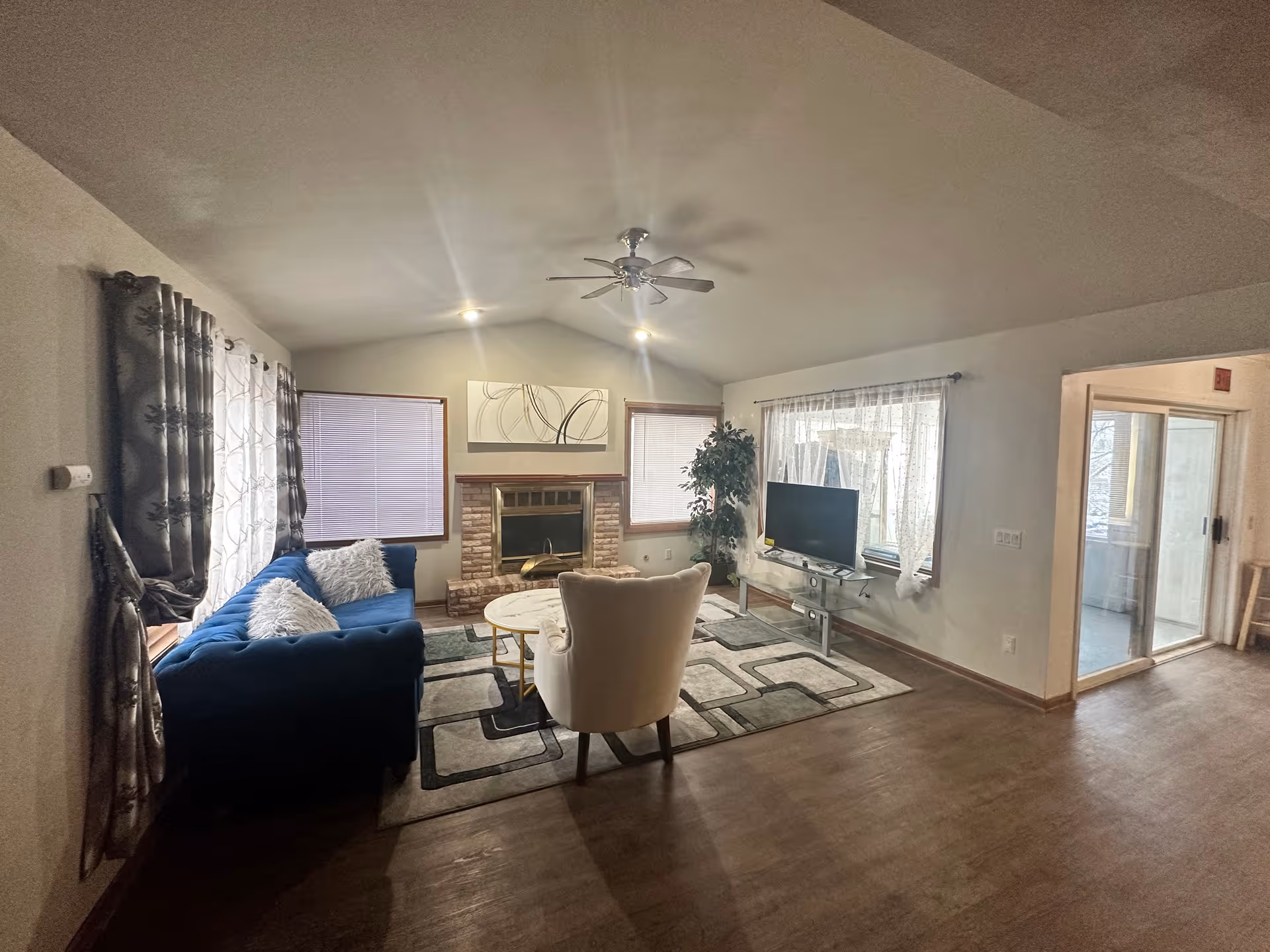 A living room with a blue couch and white armchair on a patterned rug. There is a round coffee table between the seating. The room has a brick fireplace with abstract artwork above it, two windows with blinds on either side of the fireplace, and a TV on a glass stand in front of a window with sheer curtains. A ceiling fan with lights is mounted on the ceiling. The floor is wooden, and there is an open doorway leading to a room with sliding glass doors.
