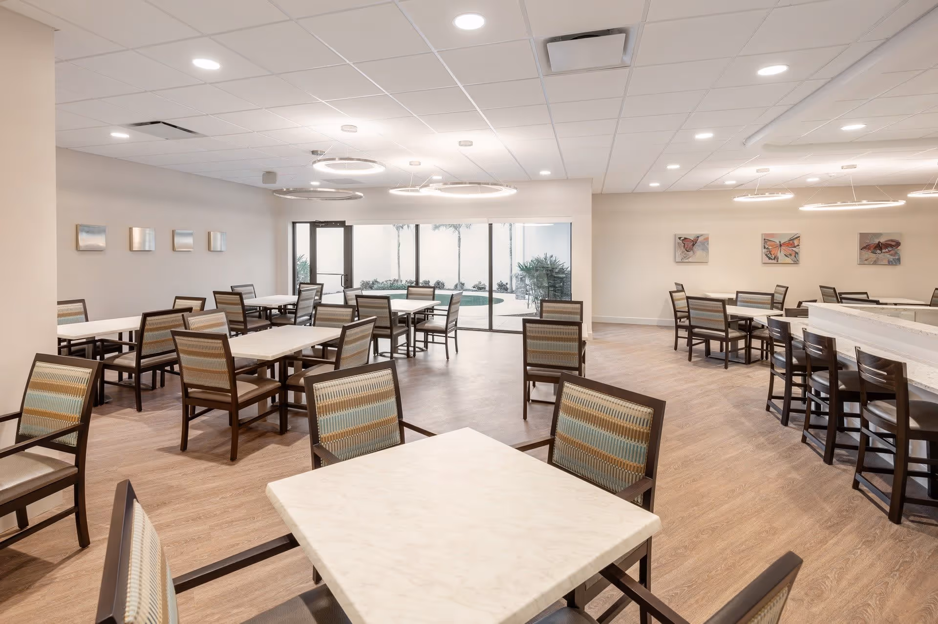 A spacious dining room with multiple tables and chairs arranged neatly. The room features light-colored walls, wood flooring, modern circular ceiling lights, and large glass doors leading to an outdoor area with plants and palm trees. The walls are decorated with small framed artworks.