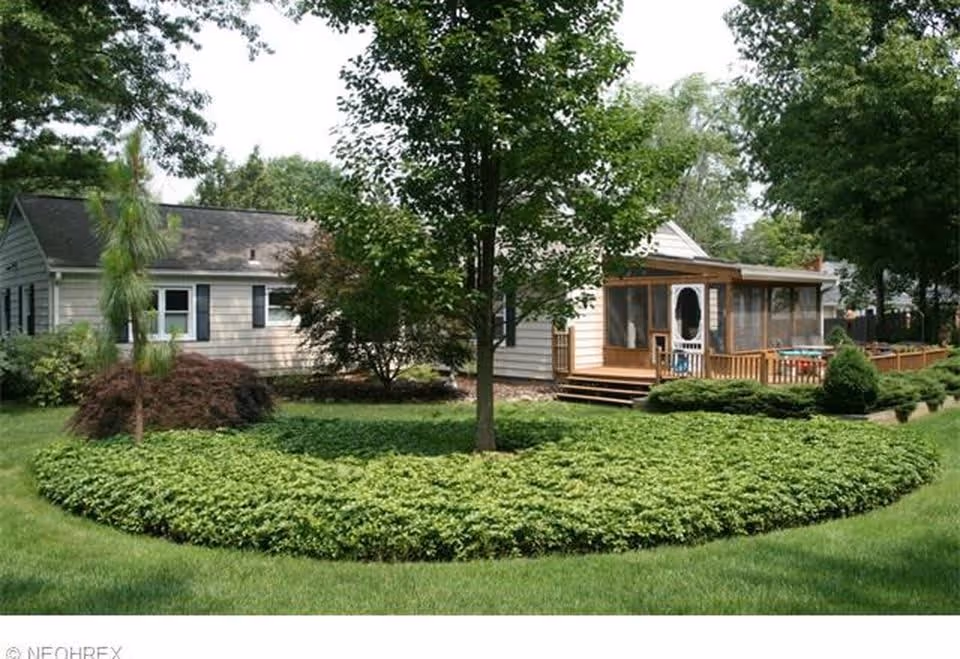 Exterior view of a single-story house with white siding and black shutters, surrounded by green trees, bushes, and a well-maintained lawn with a circular hedge in the foreground.