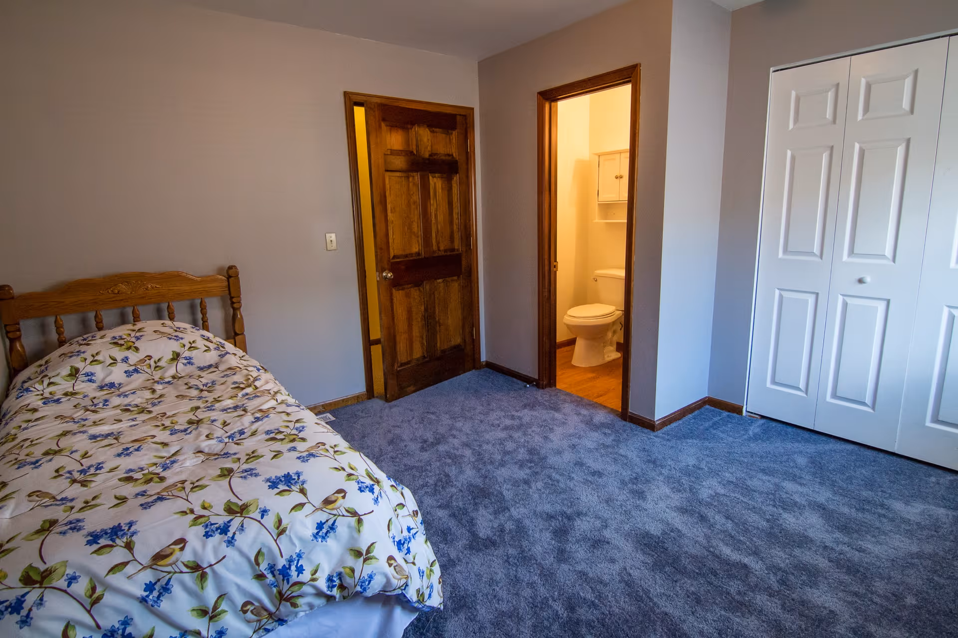 A bedroom with a wooden bed frame and floral patterned bedding. The room has blue carpet flooring, beige walls, a wooden door, a white closet with double doors, and an open doorway leading to a bathroom with a visible toilet and a cabinet above it.